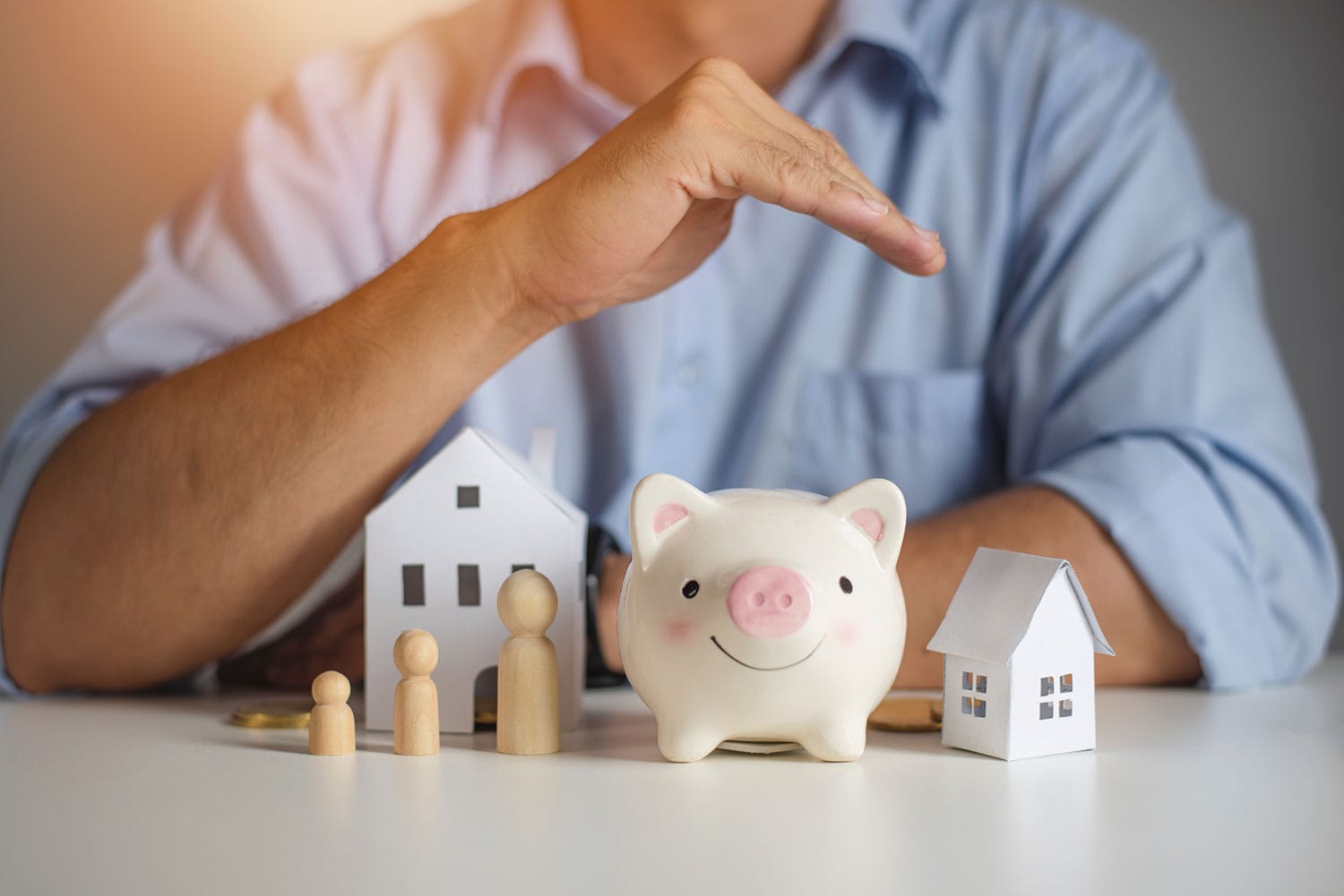 A man holding his hand slanted, like a roof over a piggy bank, wooden people and a paper house.